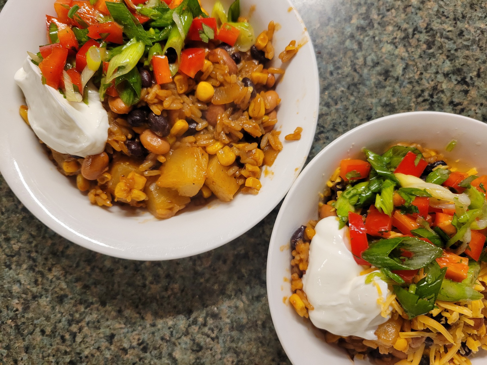 Two bowls of beans and rice with corn and pineapple. They are topped with sour cream and a bright red pepper-and-green-onion salad