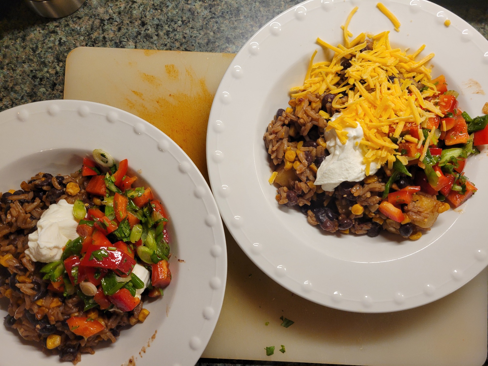 Two bowls of beans and rice, topped with sour cream and a salad of red peppers, cilantro, green onion (also one has cheese)