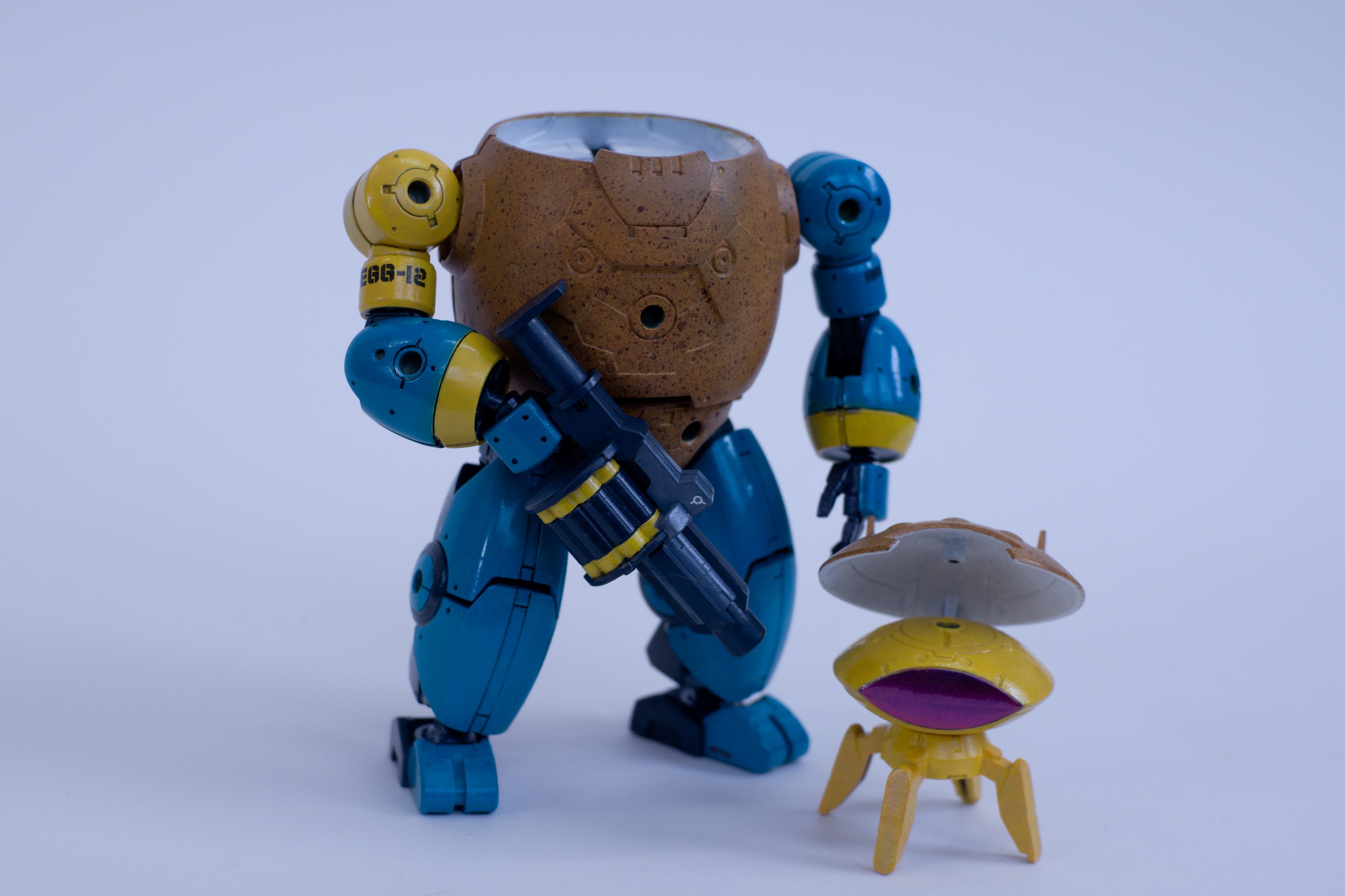A portrait of EGG-12, standing with its grenade launcher at rest in front of its body. The mini robot stands next to it, with its eggshell 'helmet' lifted up. The mini robot is egg-yolk yellow with a shiny pink-to-purple visor eye