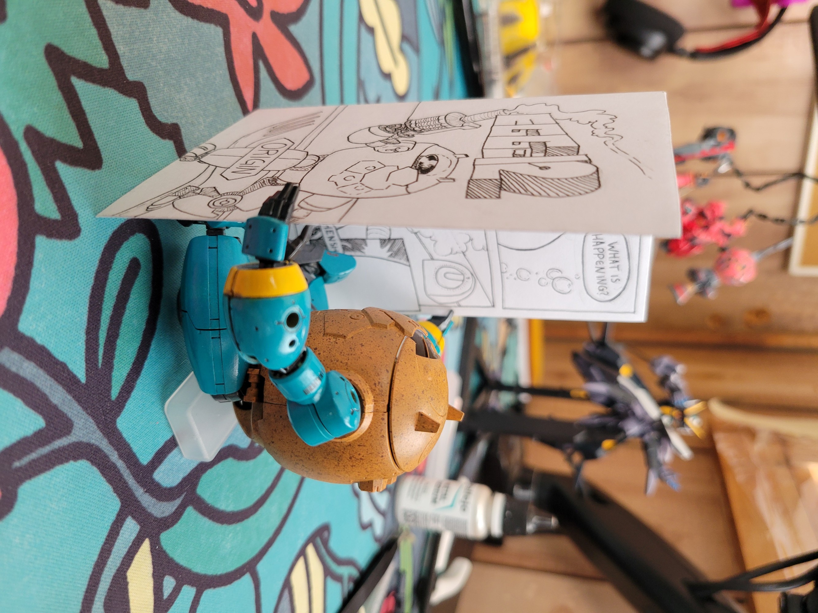 Egg 12 sits on a computer desk mat, reading a zine about itself. The zine is larger than the robot.