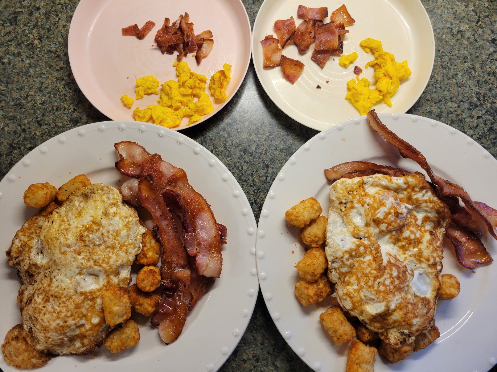 Two large plates and two plastic plates. The lare plates have bacon and tatertots covered by overeasy eggs, well fried. The small plastic plates have scrambled eggs and bacon cut into bite-sized pieces