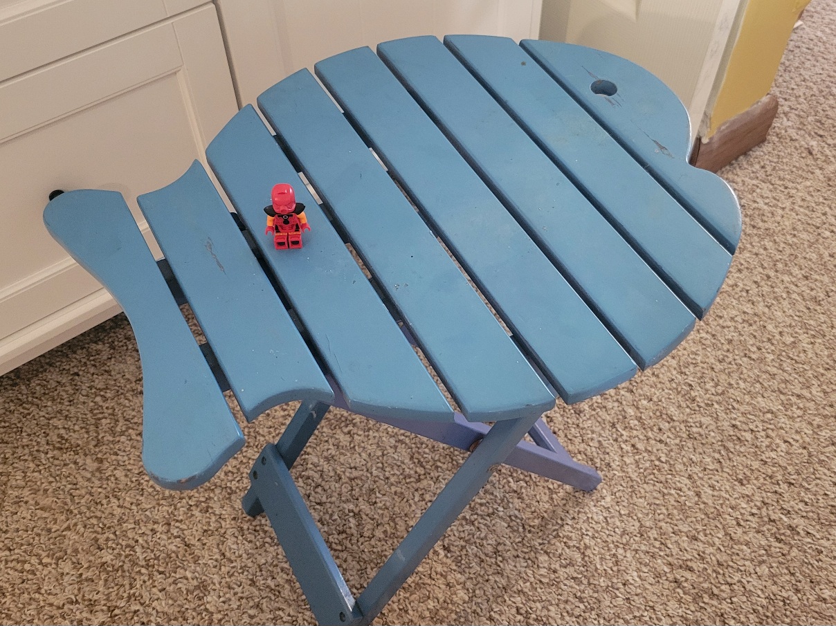 a blue folding table with a Lego minifig sitting on it for scale. The tabletop is cut to the shape of a fish and slatted, making it look like a cartoon skeleton fish
