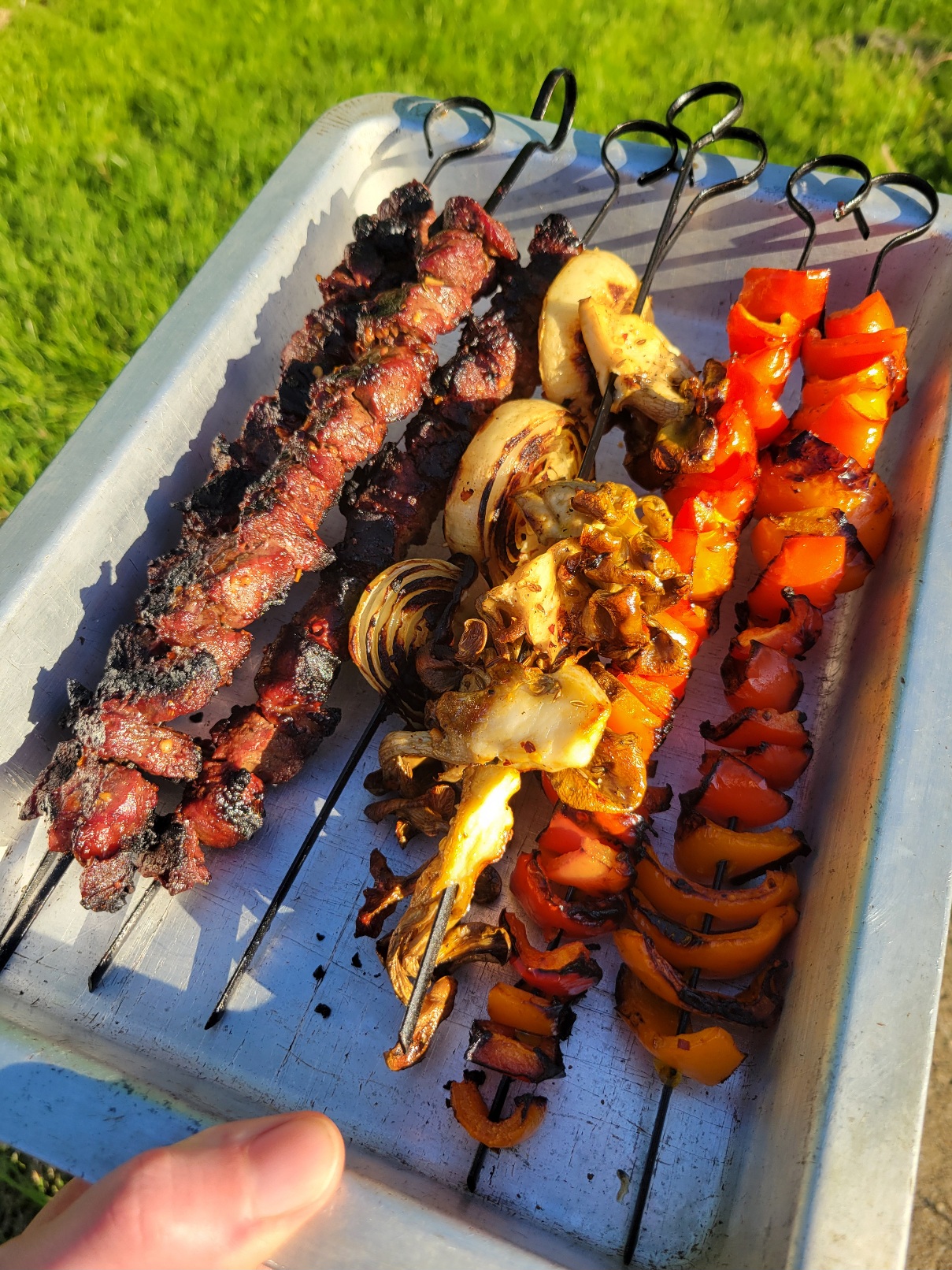 A metal pan holding skewers of steak, peppers, onions, and mushrooms. Each skewer has only one type of food on it and are all beautifully charred