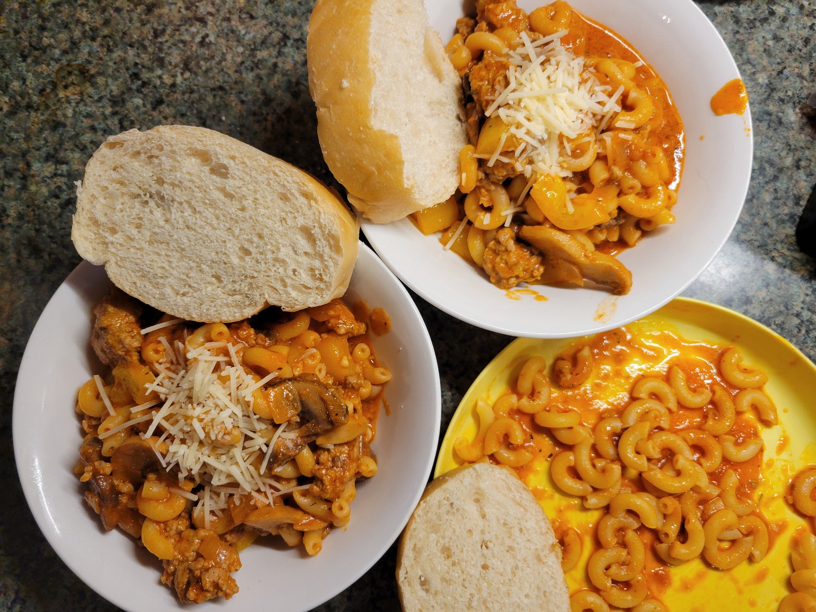 Two bowls and a plate of pasta in a creamy red sauce. Each vessel has a slice of bread; the plate noteably is only noodles and sauce, no mushrooms, sausage, or peppers.