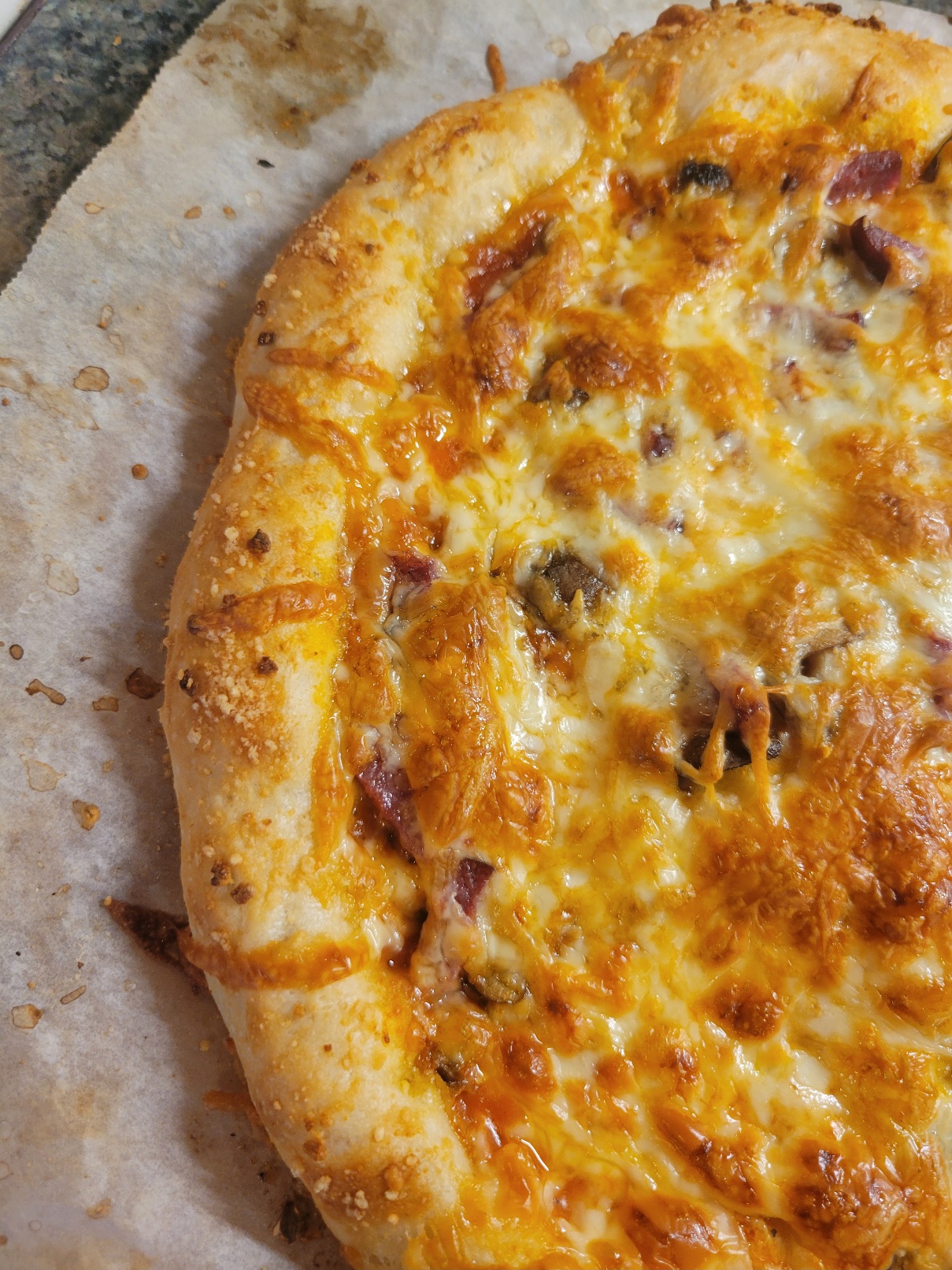 A pizza, uncut, fills the frame, its crust desribing an arc from edge to edge. The cheese is golden brown, toppings just barely peeking out from underneath