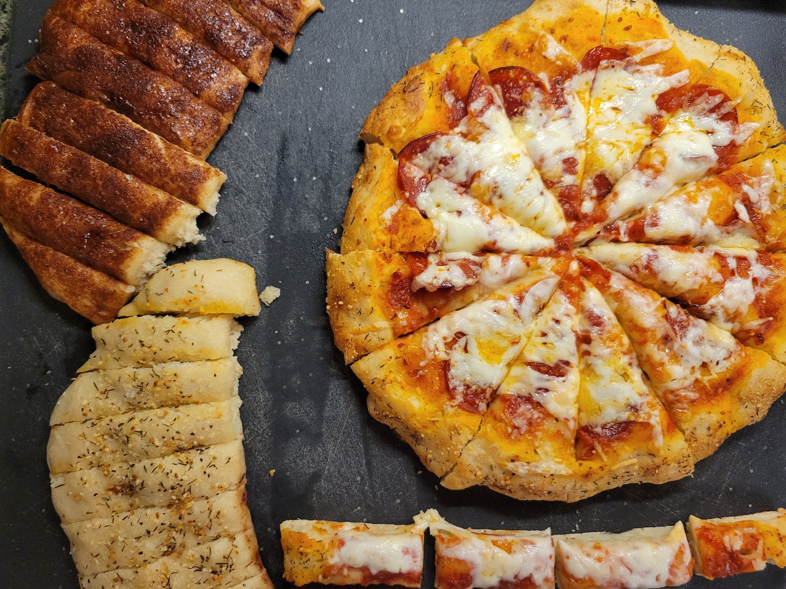 A peperoni pizza on a cuttingboard with cinnamon breadsticks,  lemon pepper breadsticks, and some cheese pizza shapped like breadsticks