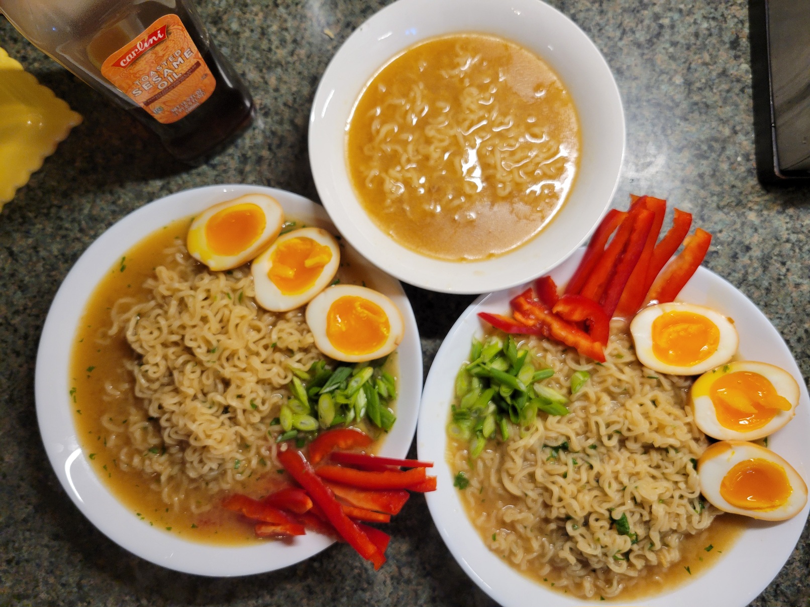 Three bowls of ramen, one small and plain, two topped with three egg halves, red peppers, and green onion
