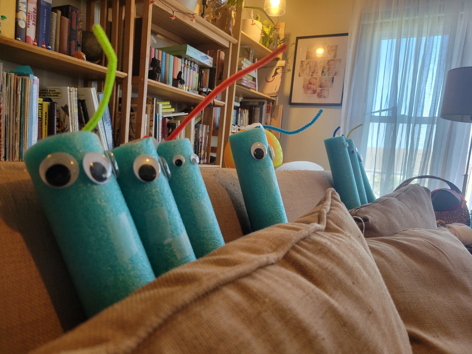Seven pool noodles with googly eyes and a single pipecleaner antenna each are shoved behind the back cushions of a couch in a stately row