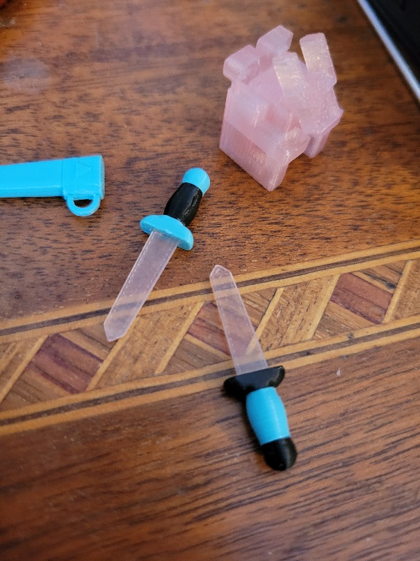 two tiny 3D printed swords with transparent blades next to a clear-ish pink cubic gargoyle