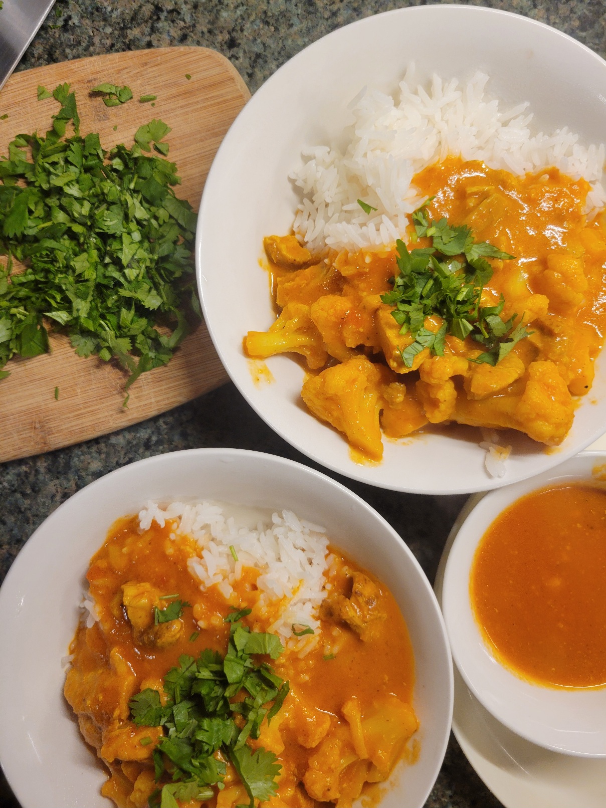 two bowls of rice and tika masala curry next to a small bowl of only the gravy. There is also part of a cutting board with chopped cilantro in frame