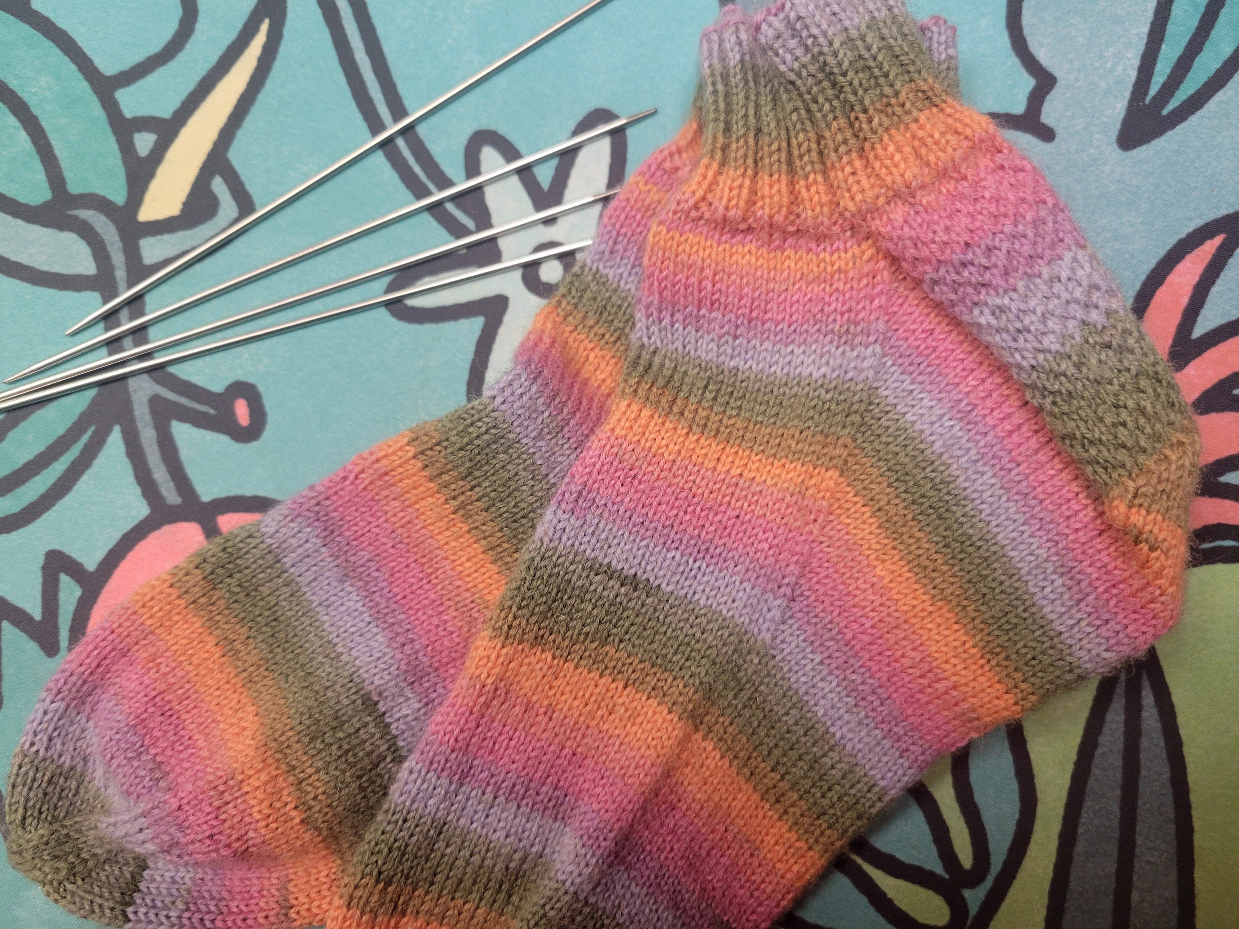 a pair of handknit socks. They have stripes of earthy green and the orange, pink, and purple of a good sunset