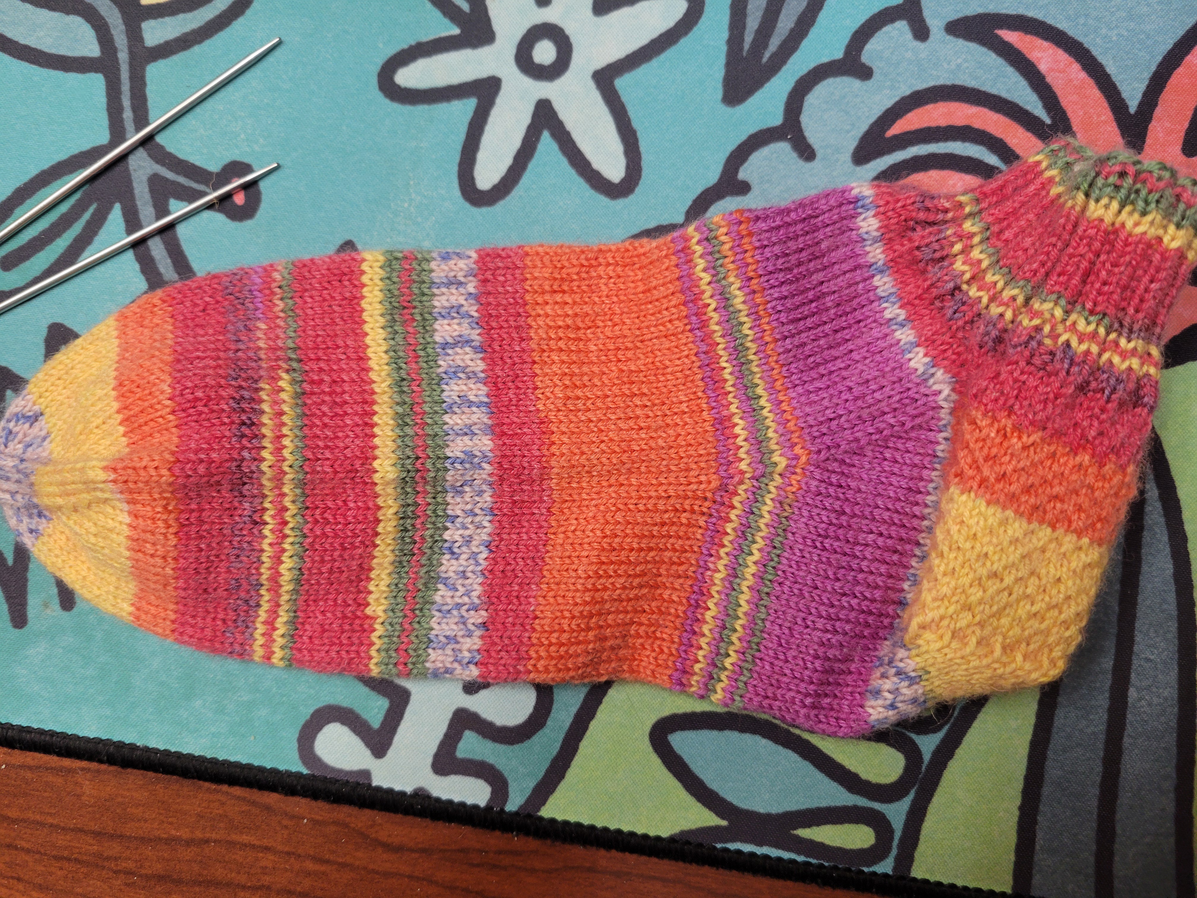 a hand knit sock with stripes of yellow, orange, pink, and green plus a stripe of mottled white and blue
