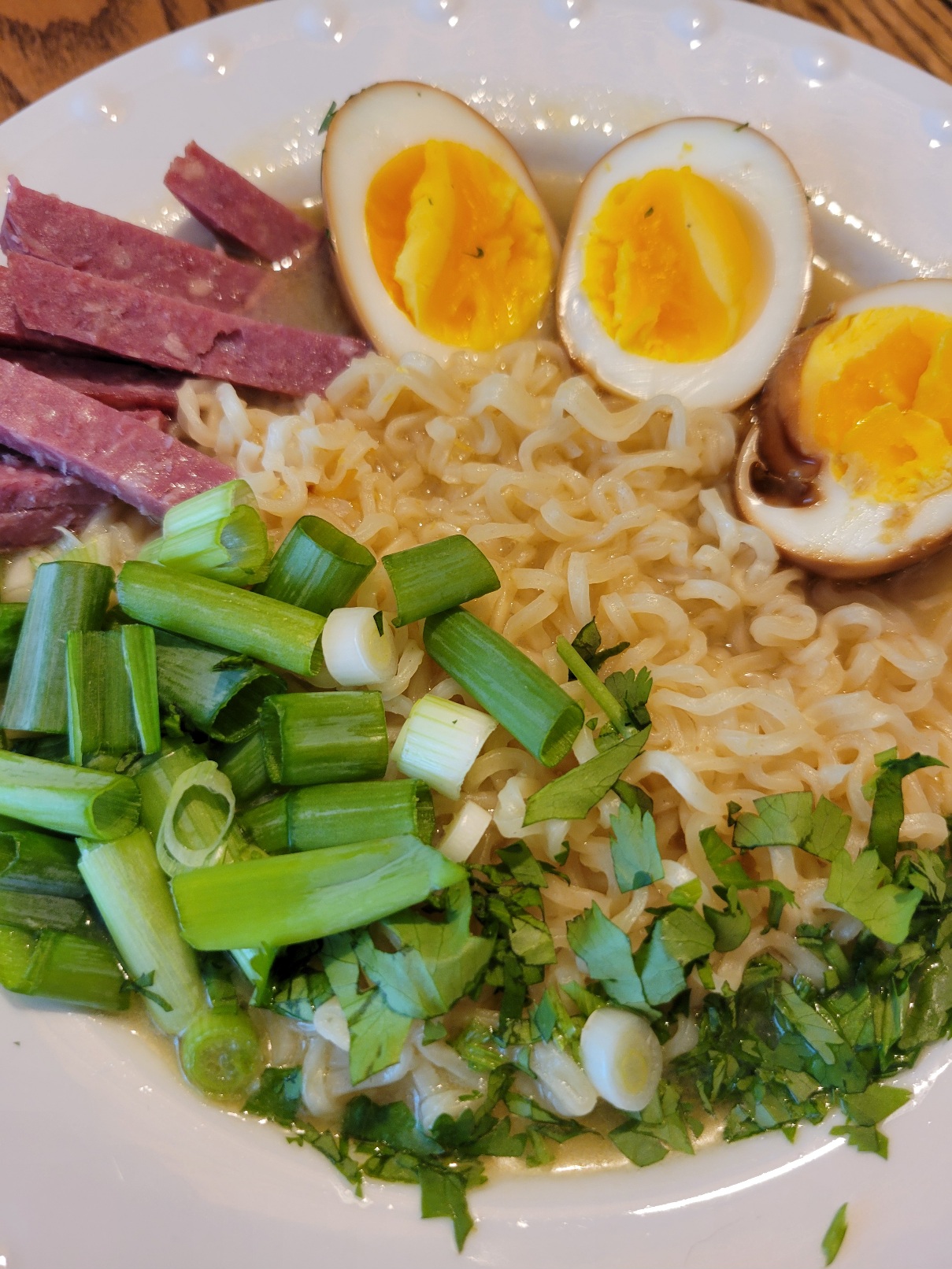 a bowl of ramen with green onion, cilantro, three brined egg halves, and salami cut into sticks