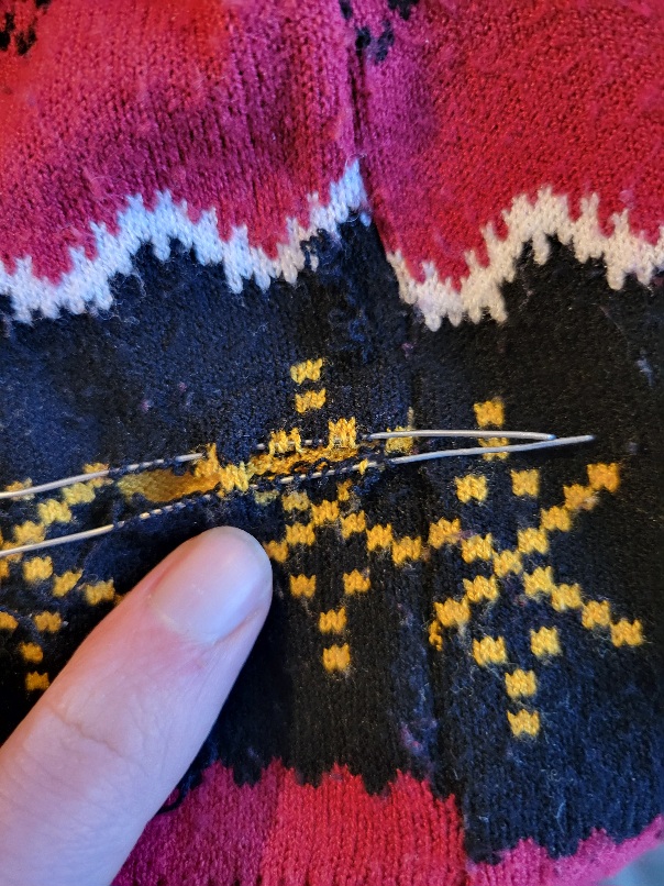 a black, yellow, and red sweater with a tear in it. The yellow underneath the black is still holding it together, but many black stitches are picked up with bits of wire