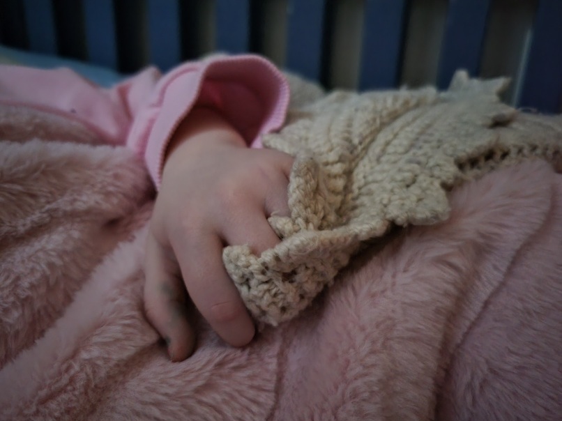 a low light photo of a child's hand. The fingers are woven into the folds of a knit blanket