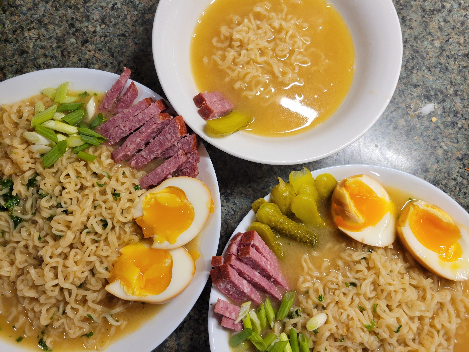 three bowls of ramen, with an arrangement of pickles, summer sausage cut into sticks, green onion, and cilantro. The smallest bowl only has two sausage sticks and a slice of pickle