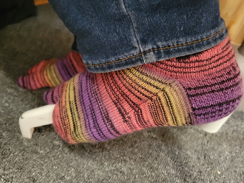 a close up of a pair of hand knit socks being worn. The socks are a gradient of purple, pink, orange, and yellow and have tabi toes