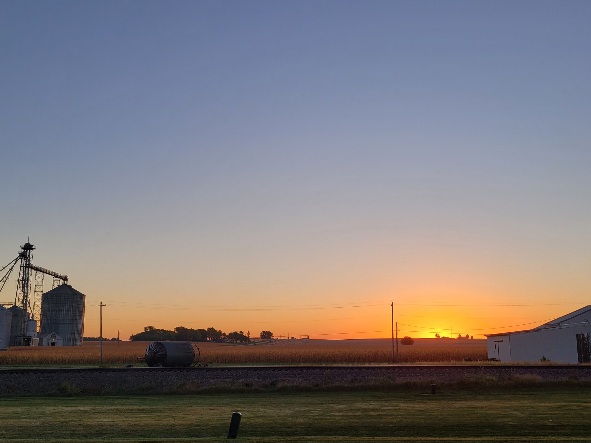 a glowing orange horizon over a wide cornfield framed by silos and a machine shed